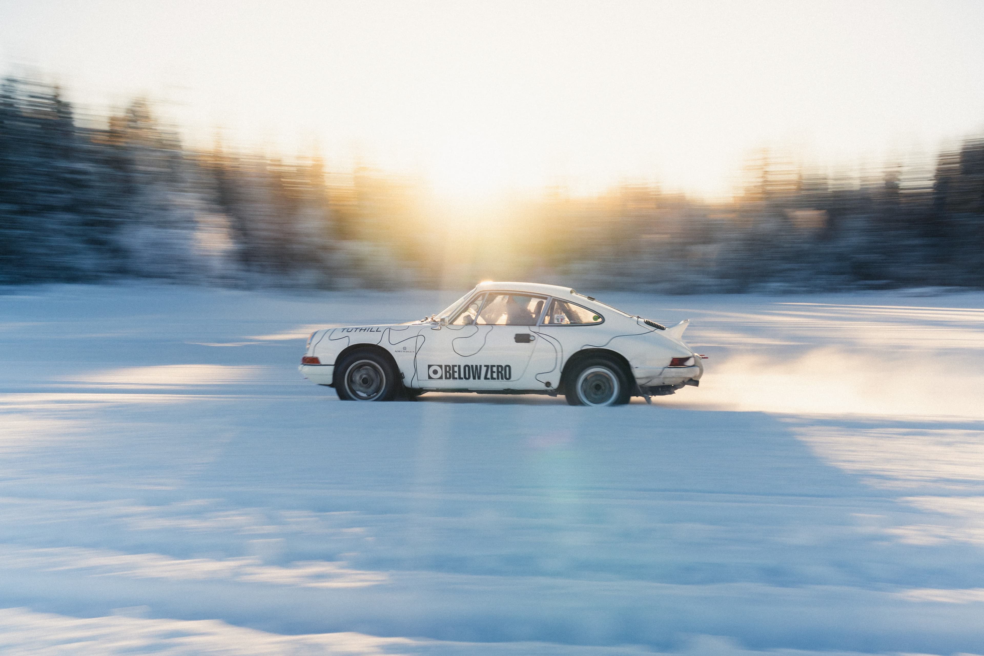 White Porsche 911 rally car drifting on a snow-covered track with a blurred forest background and sunlight streaming from behind.